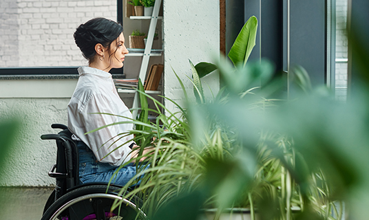 A woman works at a desk in her house. She is using a wheelchair and surrounded by plants and books.